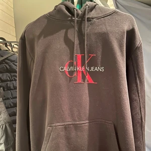 Svart hoodie från Calvin Klein Jeans - Svart hoodie från Calvin Klein Jeans med stor röd CK-logga och vit text på bröstet. Tröjan har huva med snörning och en klassisk magficka. Perfekt för dig som gillar stilrena och snygga streetwear-plagg.