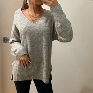 Grå stickad v-ringad tröja - Mysig grå stickad tröja med v-ringning och långa ärmar. Tröjan har en loose passform och ribbade muddar vid ärmslut och nederkant. Snygga slitsar på sidorna ger en chill vibe. Perfekt att matcha med jeans eller leggings.