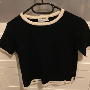 Svart stickad topp från Zara - Svart stickad topp från Zara med kontrasterande vita ribbade kanter vid halsringning, ärmslut och nederkant. Modellen är kortärmad och har en klassisk, rund hals. Perfekt för dig som gillar enkel och stilren design.