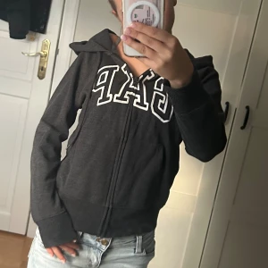 Svart GAP hoodie med dragkedja - Mysig svart hoodie från GAP med vit broderad logga över bröstet. Tröjan har huva, dragkedja framtill och fickor på sidorna. Perfekt för chill dagar eller när du vill ha en enkel och snygg look.