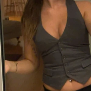 Mörkgrå omlottväst med knappar - Snygg mörkgrå väst med v-ringad hals och omlott-design. Västen har knappar framtill och två dekorativa fickor. Passformen är tight och den är croppad, vilket ger en modern och trendig look. Perfekt att styla över en t-shirt eller direkt på huden.