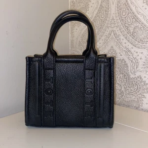 Svart handväska i skinn - Snygg svart handväska i strukturerat skinn med stilrena handtag och avtagbar axelrem med guldfärgade detaljer. Väskan har en boxig form och präglad logga på framsidan. Perfekt storlek för det viktigaste och ger en cool vibe till din outfit.