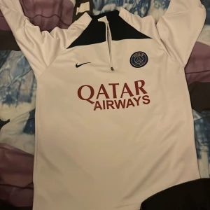 Vit PSG långärmad fotbollströja Nike - Snygg vit långärmad fotbollströja från Nike med Paris Saint-Germain-märke på bröstet och Qatar Airways-tryck. Tröjan har svart detalj vid halsen och halv dragkedja. Tillverkad i mjukt, sportigt material som andas.