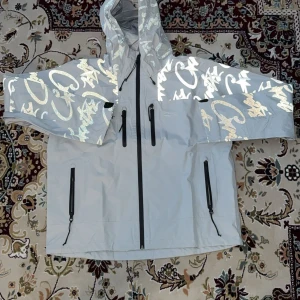 Grå vindjacka Cortez Elite med reflex - Snygg grå vindjacka från Cortez Elite med huva och feta reflexdetaljer i graffiti-stil på ärmar och axlar. Jackan har flera svarta dragkedjor framtill och coolt tryck på insidan. Perfekt för dig som vill sticka ut och hålla dig torr.