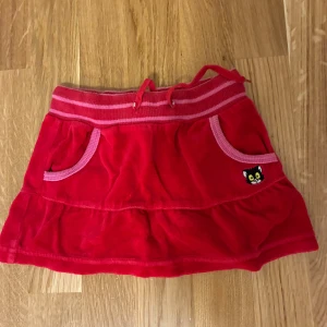 Röd Bamse-kjol från Lindex, stl 92 - Supersöt röd kortkjol från Bamse by Lindex med rosa detaljer och randig resår i midjan. Kjolen har fickor fram, en liten Bamse-katt-patch och volangkant nertill. Perfekt för dig som gillar färg och lekfull stil.