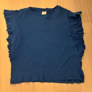 Blå ribbad topp med volang från ZARA - Säljer en blå ribbad topp från ZARA med volangdetaljer längs axlarna. Toppen är ärmlös och har en rund halsringning. Perfekt för dig som gillar en enkel men ändå cool look med extra detaljer på sidorna. Pris kan diskuteras!