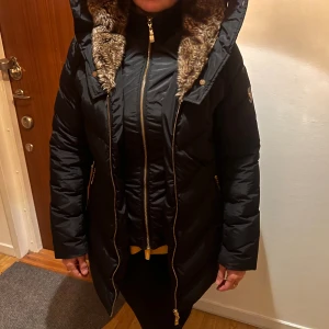 Svart lång dunjacka Hollies  - Snygg svart lång dunjacka från Hollies med guldig dragkedja och detaljer. Jackan har en stor huva fodrad med mjuk fuskpäls i brunt och två dragkedjeförsedda fickor. Perfekt för kalla dagar och riktigt stilren look.                          Tyvärr får jag inte nån användning av jackan alls, den har jag köpt förra året, användes få gånger. Storleken är 42 men sitter som 38. Priset kan diskuteras 