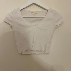 Vit ribbad topp från Pull&Bear - Säljer en vit ribbad topp från Pull&Bear i storlek XS. Toppen har v-ringning, korta ärmar och vågiga kanter vid ärmslut och nederkant. Materialet är stretchigt och är aldrig använd så mycket bra skick.
