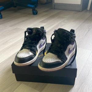Nike Air Jordan 1 Mid svart/lila - Säljer ett par Nike Air Jordan 1 Mid med svart och vitt skinn samt lila detaljer och swoosh. Klassisk high-top siluett med snörning och perforerad tå. Grymma sneakers för dig som gillar streetstyle och vill sticka ut med färg. Boxen medföljer.