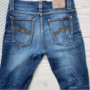 Blå jeans från Nudie Jeans - Säljer ett par blå  jeans från Nudie Jeans med klassisk femficksdesign och snygga kontrastsömmar. Jeansen har gul broderad våg på bakfickorna och är tillverkade i slitstarkt denim. Passar dig som gillar en smal siluett och cool tvätt.