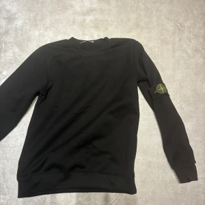 Svart Stone Island sweatshirt - Svart sweatshirt från Stone Island med klassisk rund hals och ribbade muddar. Tröjan har den ikoniska Stone Island-loggan på vänster ärm. Enkel och stilren design, perfekt för dig som gillar streetwear. Tröjan är 1:2