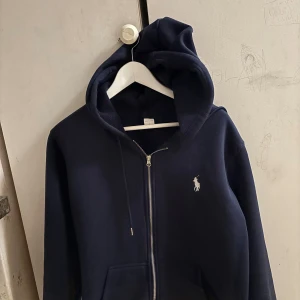Mörkblå hoodie från Polo Ralph Lauren - Mörkblå hoodie från Polo Ralph Lauren med dragkedja framtill och klassisk vit logga på bröstet. Tröjan har huva med snörning, känguruficka och är tillverkad i mjuk bomullsblandning. Perfekt för chill dagar eller när du vill ha en clean streetstyle.