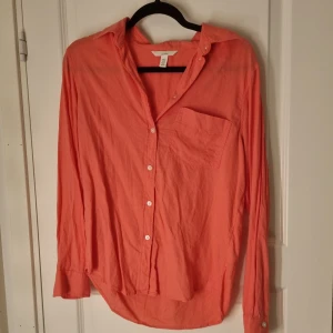 Korallrosa skjorta från H&M - Snygg korallrosa skjorta från H&M med klassisk krage och vita knappar framtill. Skjortan har en bröstficka och långa ärmar med knapp vid manschetten. Tillverkad i mjuk bomull som känns skön mot huden. Perfekt för dig som gillar färg och enkel stil.