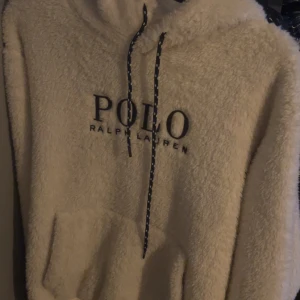 Vit teddy hoodie från Polo Ralph Lauren - Supermjuk och fluffig beige hoodie från Polo Ralph Lauren med stor broderad logga på bröstet. Tröjan har huva med snörning och en stor magficka. Perfekt för kyliga dagar när du vill vara både bekväm och snygg.