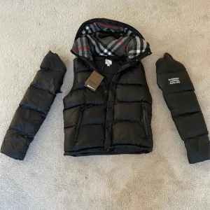 Burberry jacka/ väst - Svart vinterjacka från Burberry med avtagbara ärmar och huva. Insidan av huvan har klassiskt rutigt Burberry-mönster i grått, vitt, svart och rött. Västen har dragkedja, knappar och två fickor med dragkedja. Stor Burberry London England-logga på ena ärmen. Storlek L