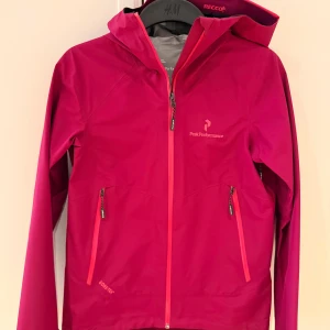 Rosa vindjacka / skidjacka Peak Performance size S - Snygg rosa vindjacka från Peak Performance med huva och dragkedjor i matchande färg. Jackan har två praktiska fickor med dragkedja och är tillverkad i slitstark polyester. Perfekt för dig som gillar att vara aktiv utomhus och vill ha en cool look. Skicket är nyskick då den är använd enstaka gånger och storleken är S