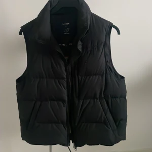 Svart dunväst från Boomerang  - Svart dunväst från Boomerang i storlek M. Västen är quiltad med hög krage och dragkedja framtill. Utsidan är svart och insidan har en beige kontrast med röd kant. Perfekt för lager-på-lager och håller dig varm under kyliga dagar. Dessa tillverkas inte heller längre och den är i stort sett oanvänd.