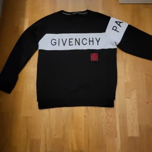 Givenchy svart sweatshirt Paris - Svart sweatshirt från Givenchy med vit panel över bröstet där det står 'GIVENCHY PARIS' i svart text. Röd Givenchy-logga broderad på framsidan. Tröjan är i 100% bomull och har långa ärmar och rund hals. Perfekt för dig som gillar streetstyle och designerplagg. De står 3XL men liten I storlek passar M/S