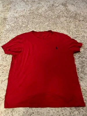 Röd t-shirt från Polo Ralph Lauren - Klassisk röd t-shirt från Polo Ralph Lauren med rund halsringning och den ikoniska lilla blå logotypen broderad på bröstet. Tillverkad i mjuk bomull och har en normal passform. Perfekt till jeans eller shorts för en clean och enkel look.