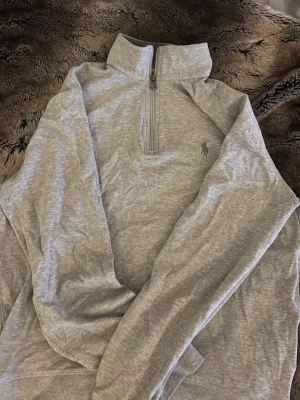 Grå halvzip från Polo Ralph Lauren - Snygg grå långärmad tröja från Polo Ralph Lauren med halvzip och hög krage. Tröjan har det klassiska broderade logotypen på bröstet och är gjord i mjukt bomullsmaterial. Perfekt för en clean och avslappnad stil.