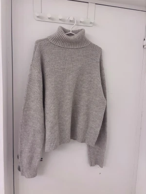 Grå polotröja i ribbstickat - Mysig grå stickad polotröja med bred ribbstickning och långa ärmar. Tröjan har en hög, vikt krage och avslappnad passform. Perfekt för kyliga dagar och enkel att matcha med jeans eller kjol.