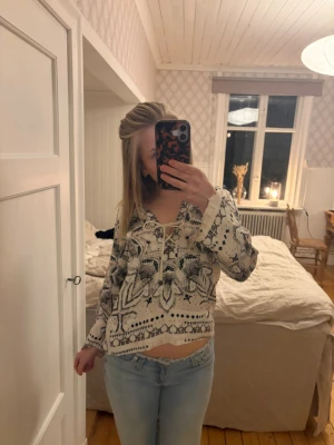 Mönstrad boho-blus med snörning - Snygg boho-inspirerad blus i vitt med svart och grått mönster. Blusen har lång ärm och v-ringning med snörning framtill. Lätt och luftig modell som passar perfekt till jeans. Materialet känns mjukt och bekvämt mot huden.