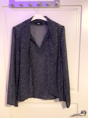 Svartprickig blus från H&M - Snygg svart blus med vita prickar från H&M. Blusen har v-ringning och långa ärmar. Tunt, lätt och luftigt material som ger en avslappnad känsla. Perfekt att styla med jeans eller kjol för en trendig look.