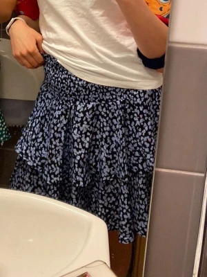 Svart volangkjol med blommigt mönster - Kort svart kjol med vita blommor och volangdetaljer. Kjolen har resår i midjan för skön passform och är gjord i ett lätt, luftigt material som passar perfekt till varmare dagar. Snygg och trendig modell som är enkel att matcha.