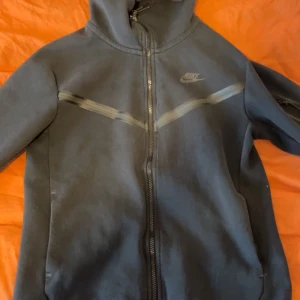 Svart Nike hoodie med dragkedja - Säljer en svart hoodie från Nike med hel dragkedja framtill och huva. Hoodien har en diskret Nike-logga på bröstet och ett grått mönster över bröstet. Perfekt för en sportig och avslappnad stil.