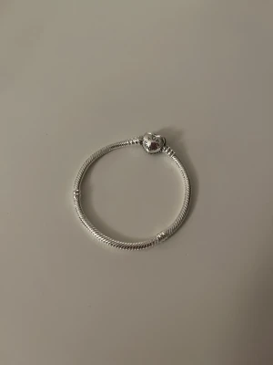 Pandora silverarmband med hjärtformad lås - Säljer ett stilrent silverarmband från Pandora med ett hjärtformat lås dekorerat med gnistrande stenar. Armbandet har en klassisk orm-länk design och passar perfekt för att addera egna berlocker. I princip nyskick!💕