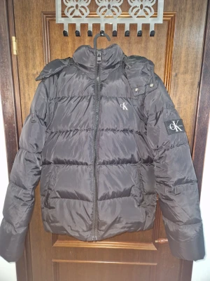 Svart pufferjacka från Calvin Klein - Svart pufferjacka från Calvin Klein Jeans med huva och dragkedja framtill. Jackan har logga på bröstet och ärmen, samt två fickor med. Perfekt för kalla dagar och har en klassisk, quiltad look.