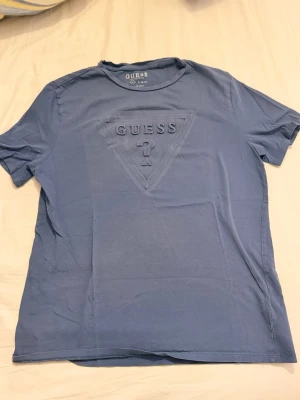  Blå t-shirt från Guess med logga - Snygg blå t-shirt från Guess med det klassiska triangelloggan och texten i relief på bröstet. T-shirten har rund halsringning och korta ärmar. Tillverkad i mjuk bomull för en skön känsla och avslappnad passform.