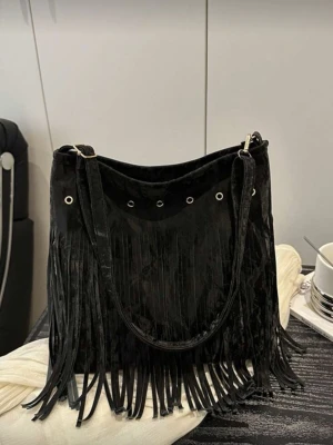 Svart axelväska med fransar - Snygg svart axelväska i mocka-look med långa fransar framtill och silverfärgade metalldetaljer. Väskan har bred axelrem och öljett-detaljer upptill för en cool boho-vibe. Perfekt för dig som vill sticka ut lite extra.