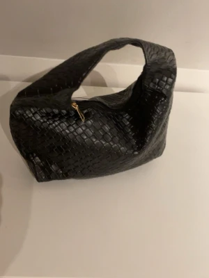 Svart flätad handväska i skinnimitation - Trendig svart handväska med flätad design och bred axelrem. Väskan har en mjuk, rundad form och stängs med en guldfärgad dragkedja. Perfekt accessoar för att lyfta din outfit.