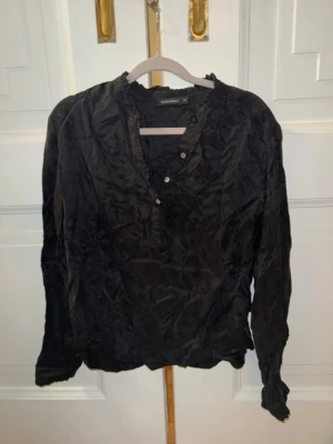 Svart blus från Rut & Circle - Snygg svart blus från Rut & Circle med lång ärm och dekorativa knappar vid halsringningen. Blusen har en lätt glansig yta och smala manschetter med små knappar. Perfekt till både jeans och kjol för en trendig look.