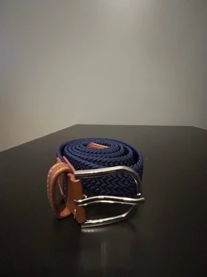 Mörkblått flätat bälte - Snyggt mörkblått flätat bälte med bruna läderdetaljer och silvrigt spänne. Helt nytt aldrig använt. Storlek 100 cm. 
