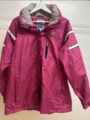 Mörkrosa vindjacka med reflexdetaljer - Rosa jacka med grå fleecefodrad krage och vita detaljer på ärmarna. Jackan har dragkedja och tryckknappar framtill, samt flera fickor med dragkedja. Perfekt vid mildare väder eller med fleece / tjocktröja under. 