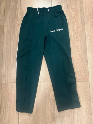 Palm Angels gröna mjukisbyxor - Mörkgröna mjukisbyxor från Palm Angels med vit snörning i midjan och vit logga på vänster sida. Byxorna har vita ränder längs sidorna och elastisk midja för skön passform. Perfekta för en chill och sportig look.