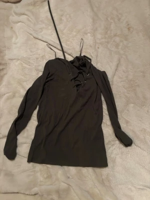 Svart ribbad långärmad topp med snörning - Trendig svart ribbad topp med lång ärm och snörning framtill i halsen. Toppen har en smickrande passform och är perfekt för att lyfta vilken outfit som helst. Snygga detaljer med snörning ger en edgy känsla.