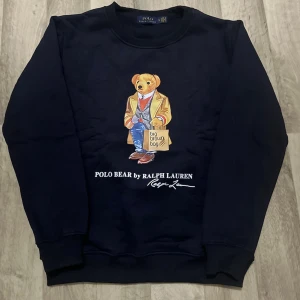 Mörkblå sweatshirt Polo Bear Ralph Lauren - Mörkblå sweatshirt från Ralph Lauren med ikoniska Polo Bear-trycket på bröstet. Björnen har beige kavaj, jeans och röd tröja. Rund halsringning och ribbade muddar vid ärmslut och nederkant. Perfekt för en avslappnad och trendig stil.