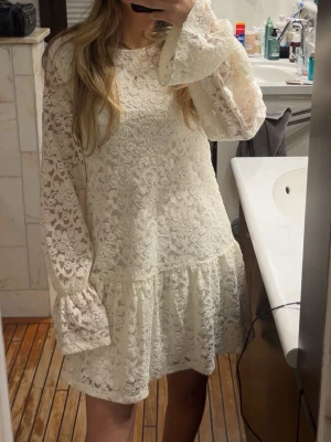 Vit spetsklänning med volang - Söt vit kortklänning i spets med volangdetalj nedtill och långa, lätt utsvängda ärmar. Klänningen har rund halsringning och ett romantiskt uttryck. Perfekt för dig som gillar en feminin och trendig stil.