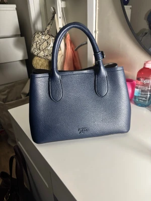 Mörkblå handväska från Ralph Lauren - Snygg mörkblå handväska. Väskan har två rejäla handtag, guldfärgade detaljer och ett rymligt beige innerfack. Perfekt storlek för det viktigaste och stilren design.