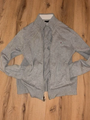 Grå kofta från Tommy Hilfiger - Säljer en stilren grå kofta från Tommy Hilfiger med dragkedja framtill och diskret logga på bröstet. Tröjan har ribbade muddar och hög krage, perfekt för lager på lager under kyliga dagar.