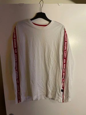 Vit långärmad Alpha Industries t-shirt - Vit långärmad t-shirt från Alpha Industries med röda band längs ärmarna där det står 'REMOVE BEFORE FLIGHT'. Rund halsringning med röd kant. Perfekt för dig som gillar streetwear och tydliga detaljer.