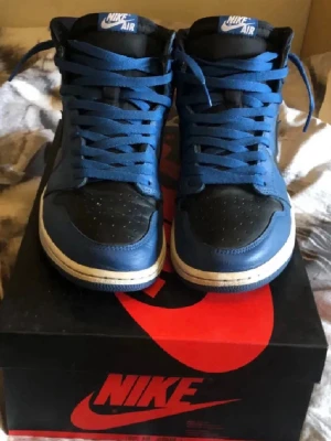 Nike Air Jordan 1 blå och svarta - Snygga Nike Air Jordan 1 sneakers i blått och svart skinn med klassisk hög siluett. Skorna har blå snörning, vit mellansula och svart yttersula. Perfekta för dig som vill ha en ikonisk och stilren sneaker med streetwear-vibe. 