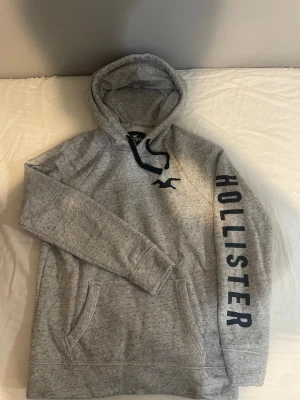 Grå hoodie från Hollister - Snygg gråmelerad hoodie från Hollister med stor huva och svart snörning. Hollister-logga tryckt längs ena ärmen och liten logga på bröstet. Mjuk och skön bomullsblandning, perfekt för chill dagar. Praktisk magficka framtill.