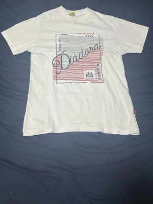Vit Diadora t-shirt med tryck - Snygg vit t-shirt från Diadora med grafiskt tryck i blått och rött framtill. Klassisk passform och rund halsringning. Tillverkad i mjuk bomull som känns skön mot huden. Perfekt för en avslappnad och stilren look.