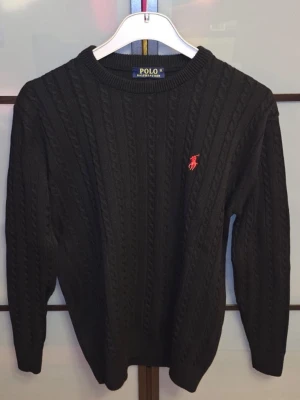 Tröja Polo Ralph Lauren - Kabelstickad svart tröja från Polo Ralph Lauren med rund halsringning och röd broderad logga på bröstet. Klassisk design med ribbade muddar vid ärmslut och nederkant. Perfekt för en stilren och tidlös look. Den är använd ett fåtal gånger. Den är i mycket bra skick. Tveka inte för att ställa frågor. Priset är inte hugget i sten.