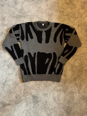 Grå stickad tröja med svart mönster - Snygg grå stickad tröja med svart abstrakt mönster. Tröjan har rund halsringning, långa ärmar och ribbade muddar vid ärmslut och nederkant. Perfekt för dig som gillar en unik och trendig look.