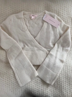 Vit omlottkofta från Gina Tricot - Söt vit stickad omlottkofta från Gina Tricot med långa ärmar och ribbad midja. Koftan har fyra små vita knappar framtill och ett band i midjan för att knyta omlotten. Perfekt till lager på lager och enkel att styla. Prislappen är kvar och den är från barnavdelningen strl 158/164